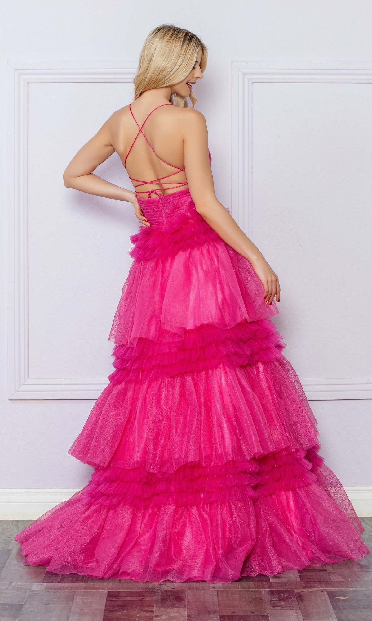 A woman, seen from behind, wears the Nox Anabel Ruffled Glitter Prom Dress R1316 in bright pink, featuring a strappy open back and ruffled details, standing before a light-paneled wall.